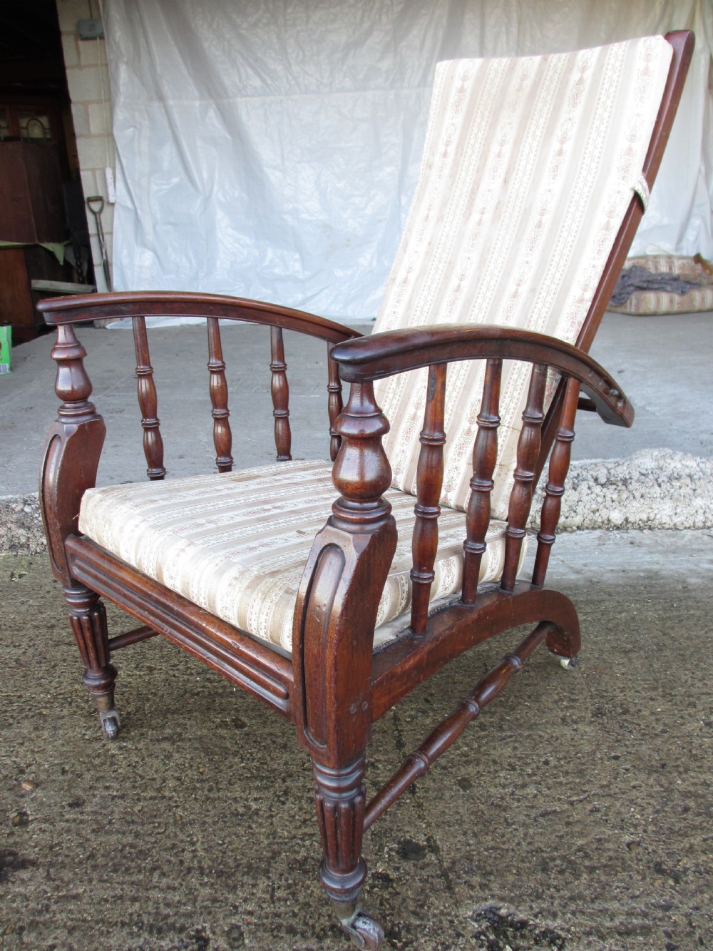 A 19th-century dark mahogany armchair featuring turned side spindles, beige striped upholstery, an adjustable reclining backrest, and brass castors.