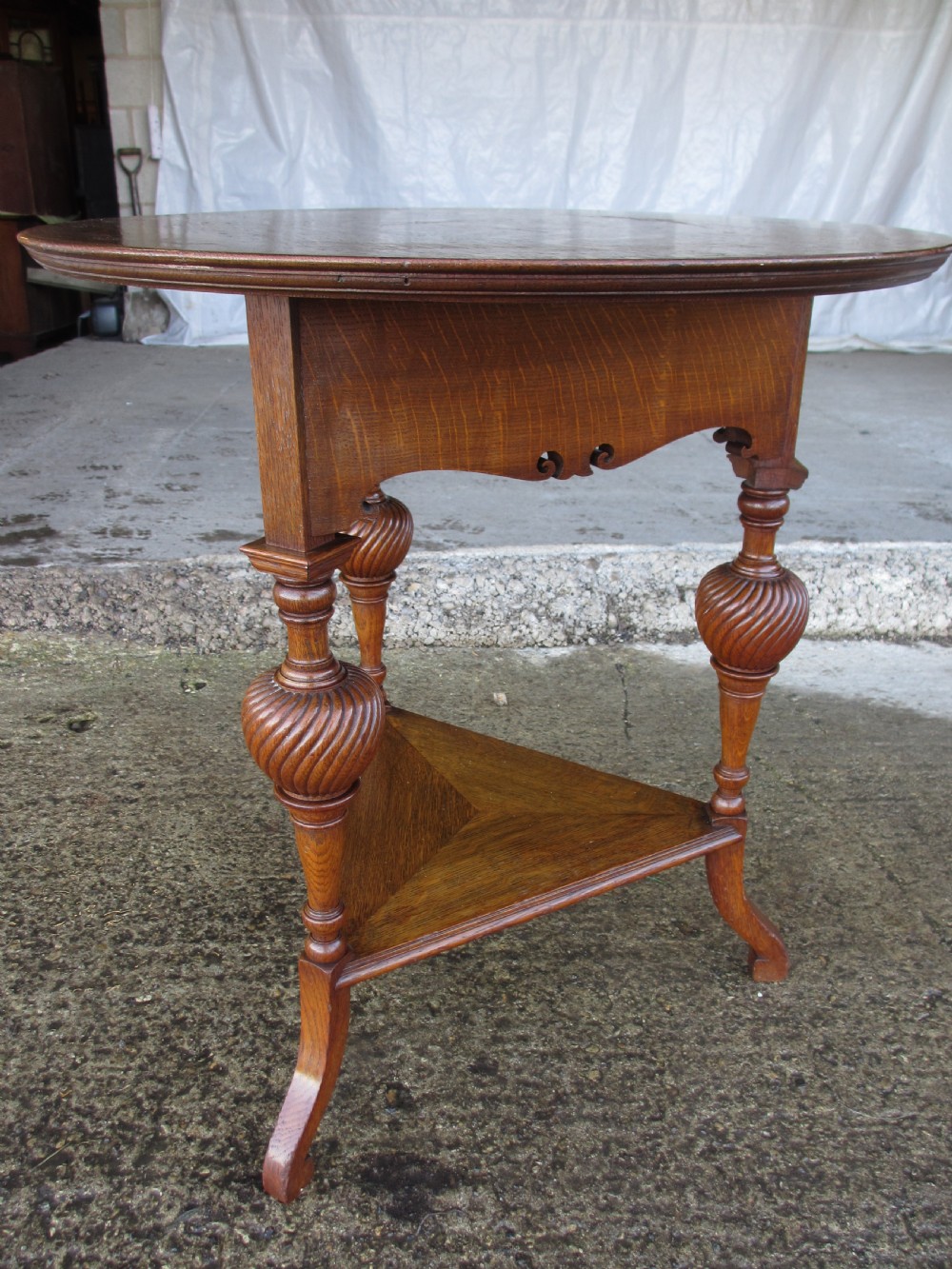 An Edwardian oak circular occasional table featuring three carved spiral-bulb legs, a shaped apron, and a lower triangular tier shelf.