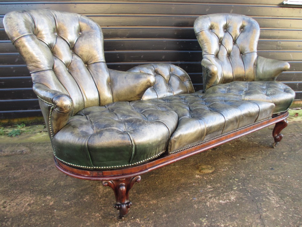 A Victorian conversation sofa upholstered in buttoned dark green leather featuring a carved rosewood frame with brass castors.