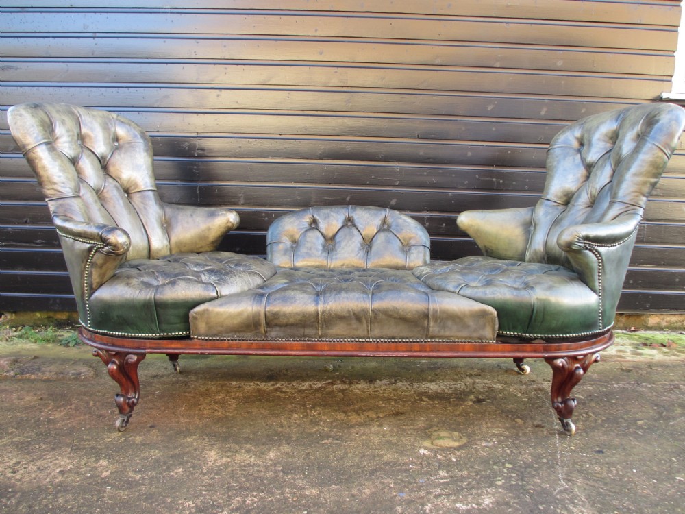 A Victorian conversation sofa upholstered in buttoned dark green leather featuring a carved rosewood frame with brass castors. View IV