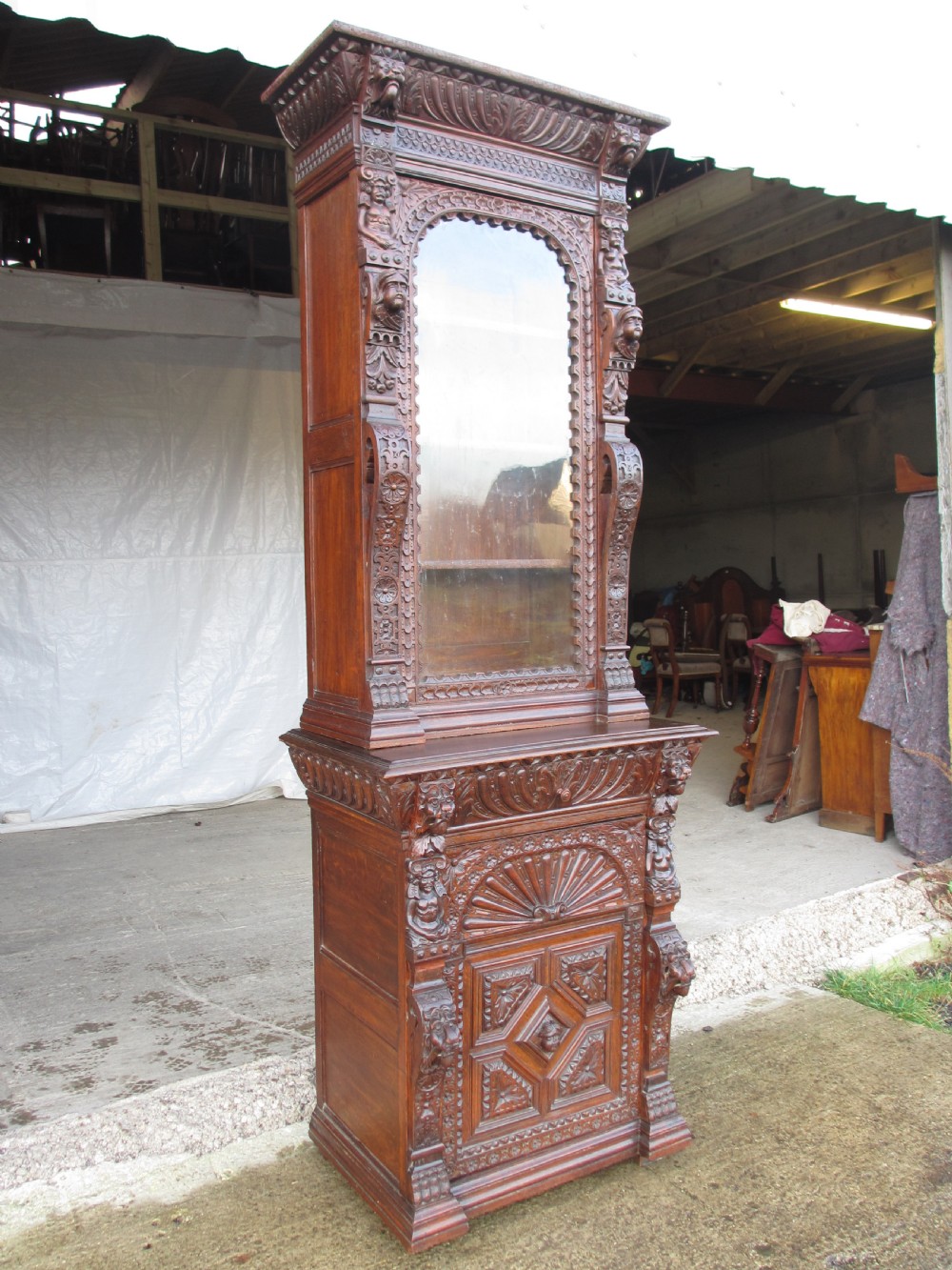 A tall two-part dark oak Victorian bookcase featuring an arched glazed upper door, carved face motifs, and geometric panelled lower drawers.