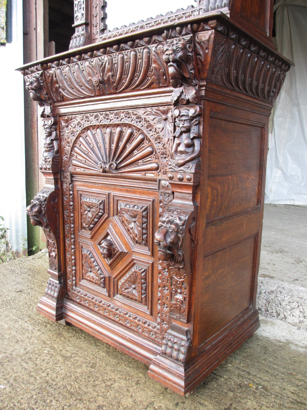 A tall two-part dark oak Victorian bookcase featuring an arched glazed upper door, carved face motifs, and geometric panelled lower drawers. View III