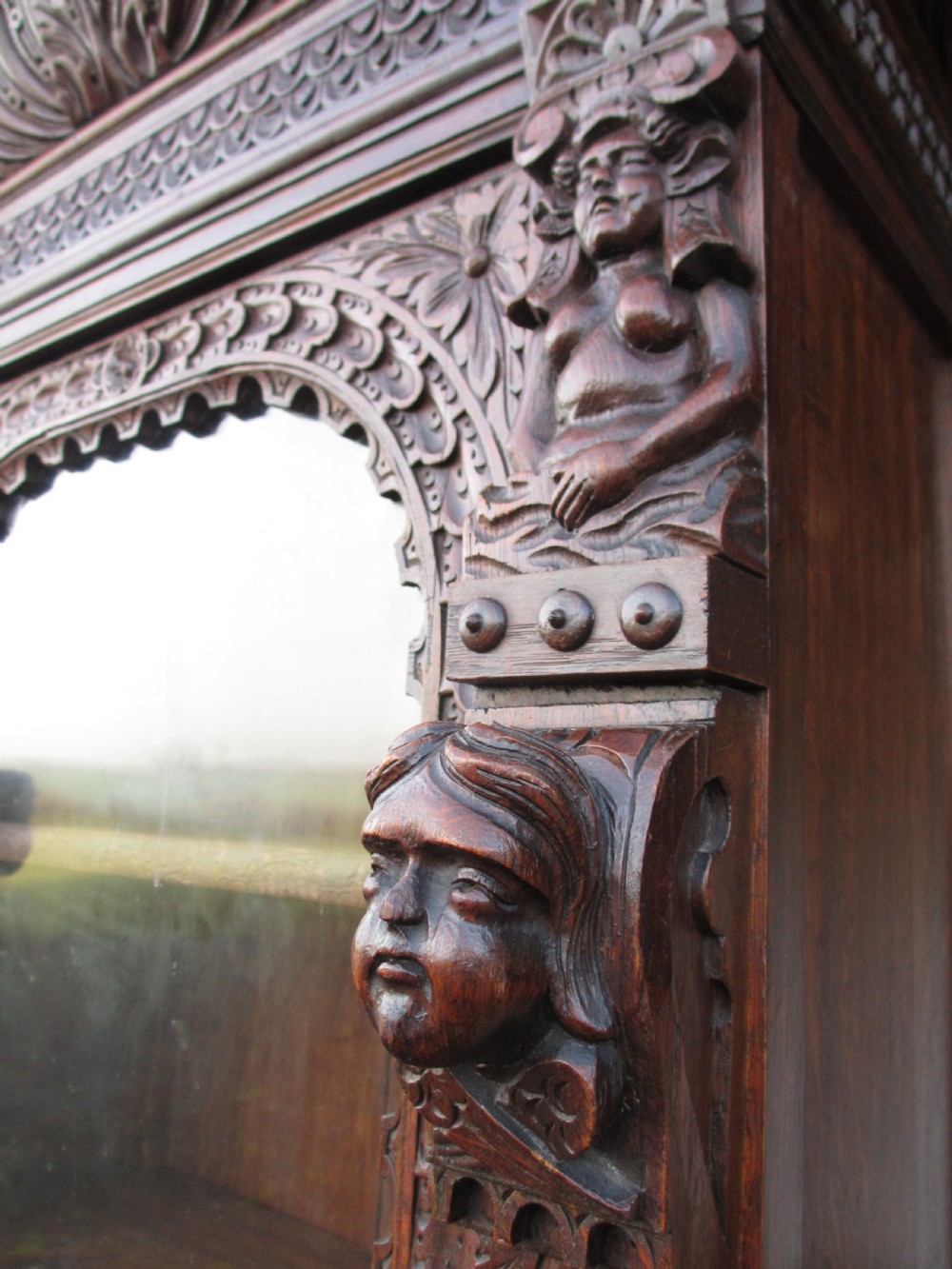 A tall two-part dark oak Victorian bookcase featuring an arched glazed upper door, carved face motifs, and geometric panelled lower drawers. View IV