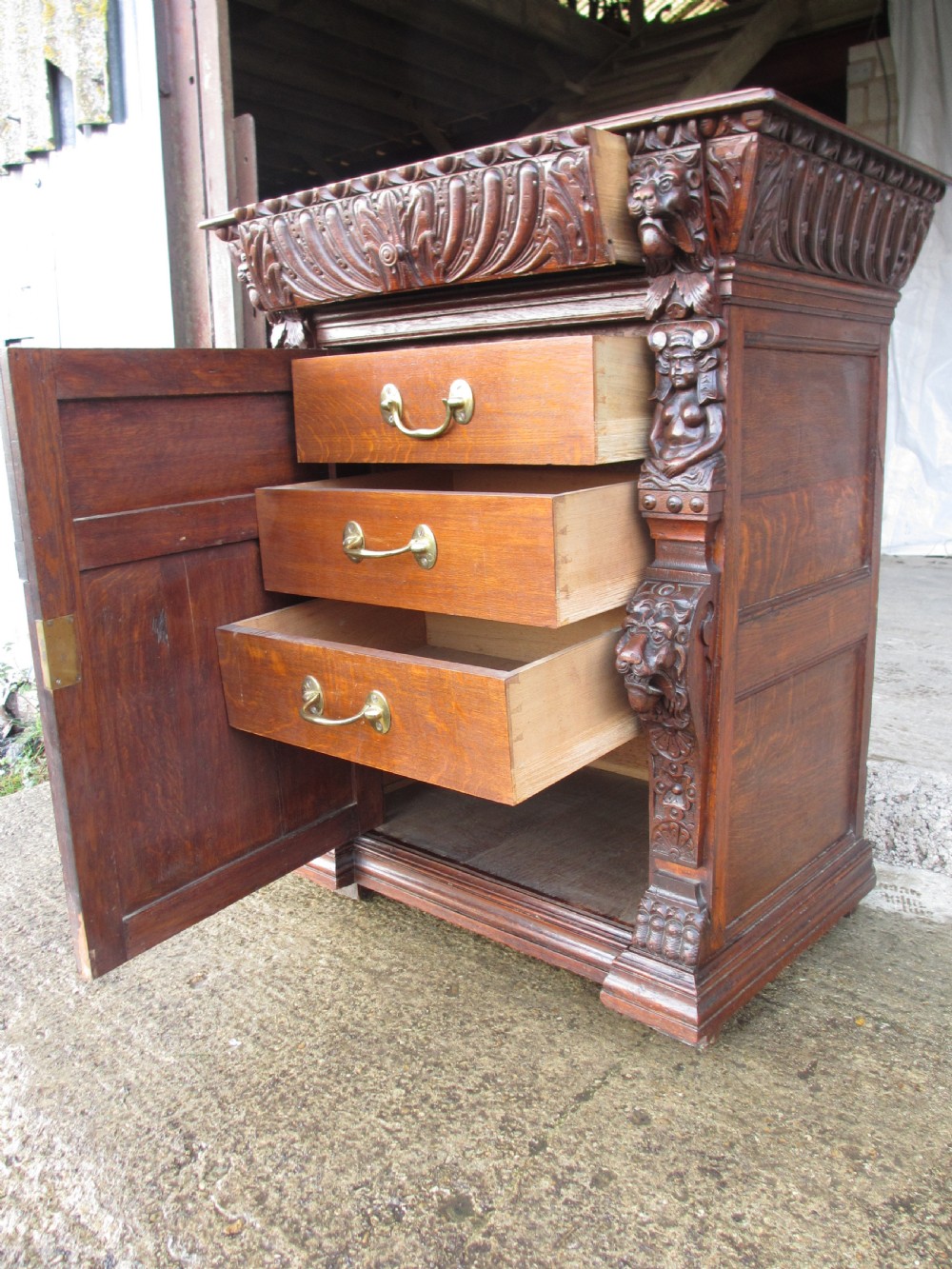 A tall two-part dark oak Victorian bookcase featuring an arched glazed upper door, carved face motifs, and geometric panelled lower drawers. View V