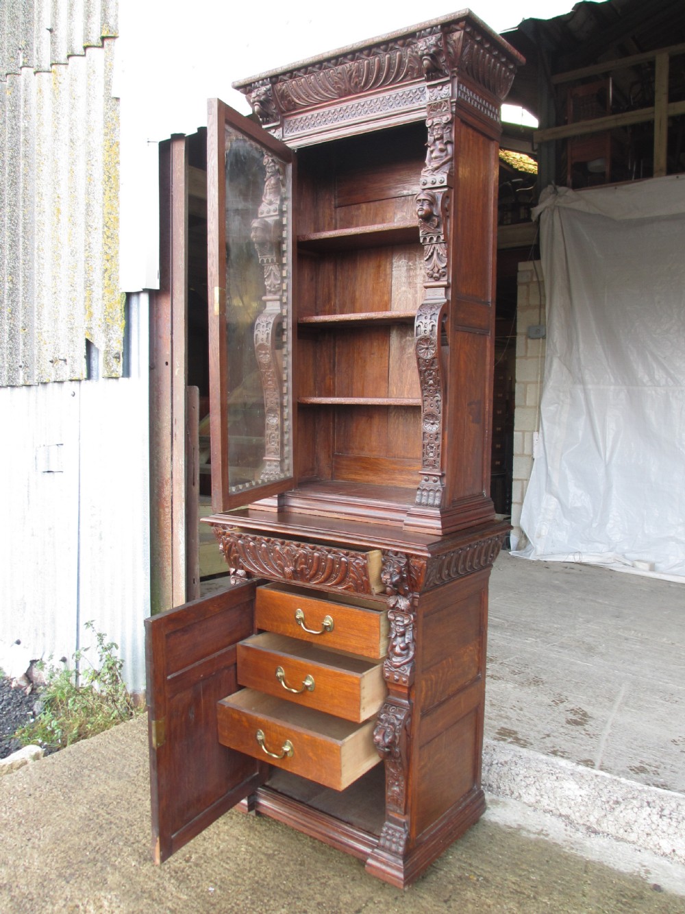 A tall two-part dark oak Victorian bookcase featuring an arched glazed upper door, carved face motifs, and geometric panelled lower drawers. View VIII