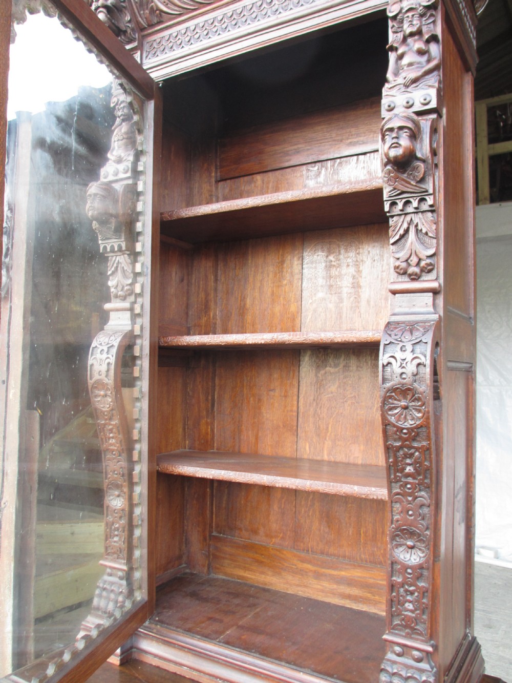 A tall two-part dark oak Victorian bookcase featuring an arched glazed upper door, carved face motifs, and geometric panelled lower drawers. View IX