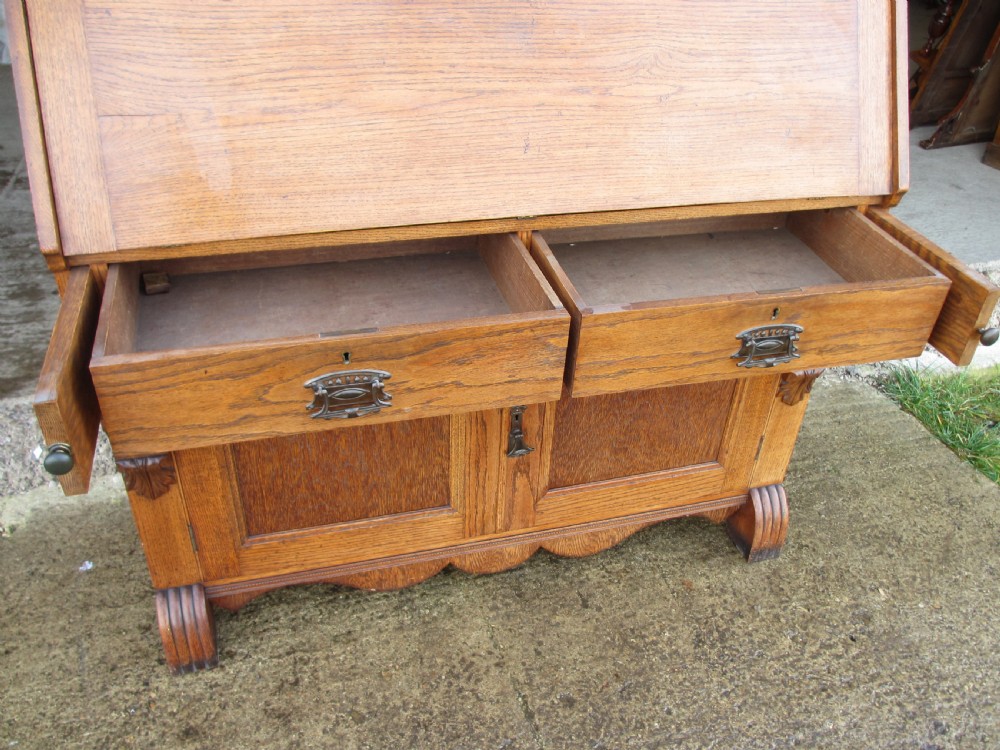 An Edwardian golden oak bureau bookcase featuring a two-door glazed upper section, a fall-front slanted writing desk, and a lower cabinet base with carved supports. View VI