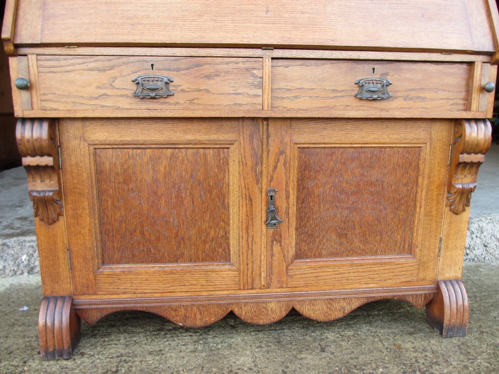 An Edwardian golden oak bureau bookcase featuring a two-door glazed upper section, a fall-front slanted writing desk, and a lower cabinet base with carved supports. View VIII