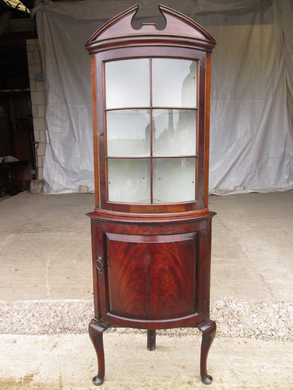 A two-piece flame mahogany corner cabinet featuring a broken pediment top, an upper glazed door, and a lower bow-front cupboard door supported by cabriole legs.