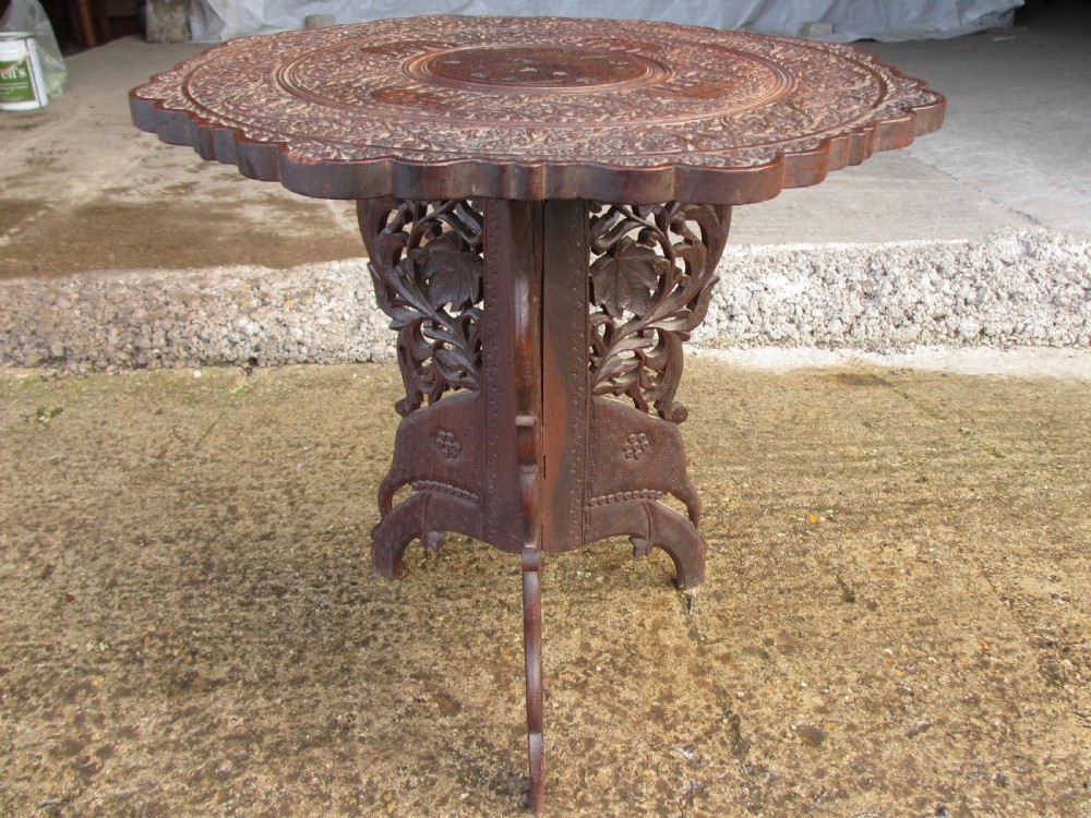 An Anglo-Indian carved teak occasional table with a scalloped circular top, floral inlay centre, and a folding fretted base. View II