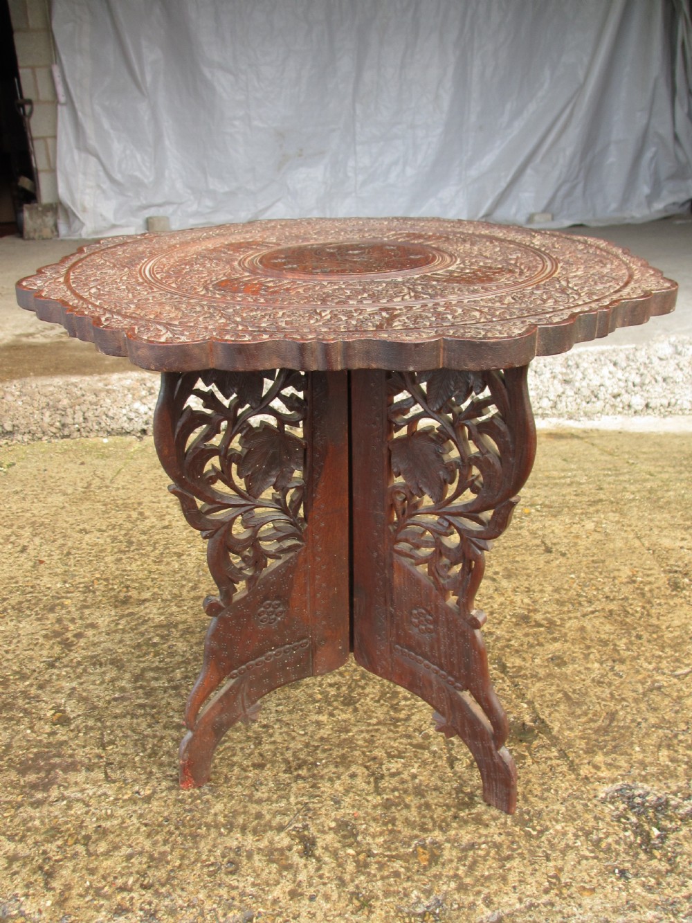 An Anglo-Indian carved teak occasional table with a scalloped circular top, floral inlay centre, and a folding fretted base. View X