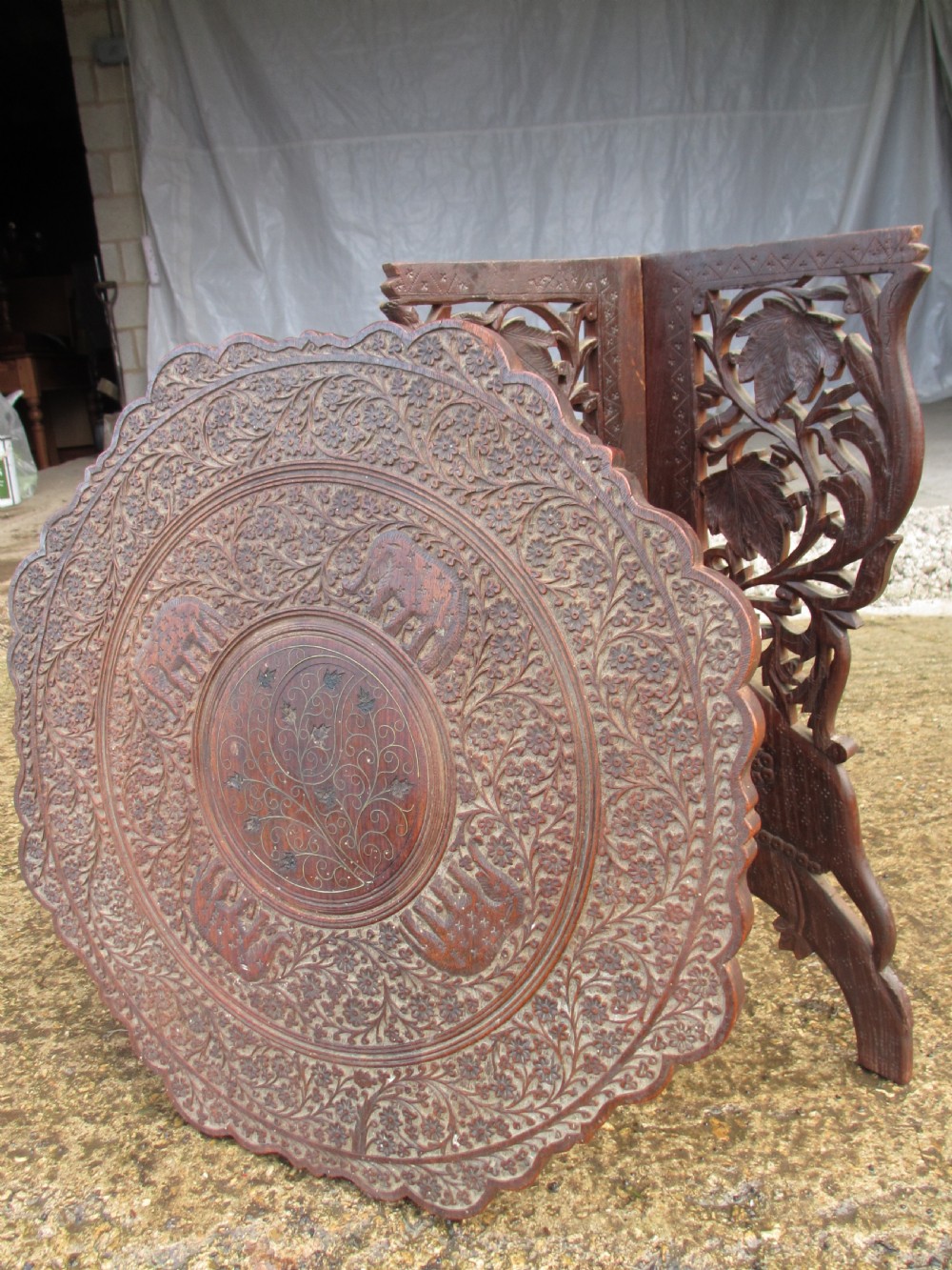 An Anglo-Indian carved teak occasional table with a scalloped circular top, floral inlay centre, and a folding fretted base. View XI