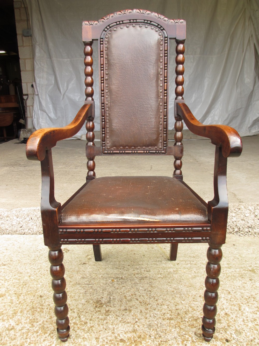 An Arts and Crafts mahogany carver armchair featuring bobbin-turned legs, curved armrests, a carved top crest, and brown studded leather upholstery.