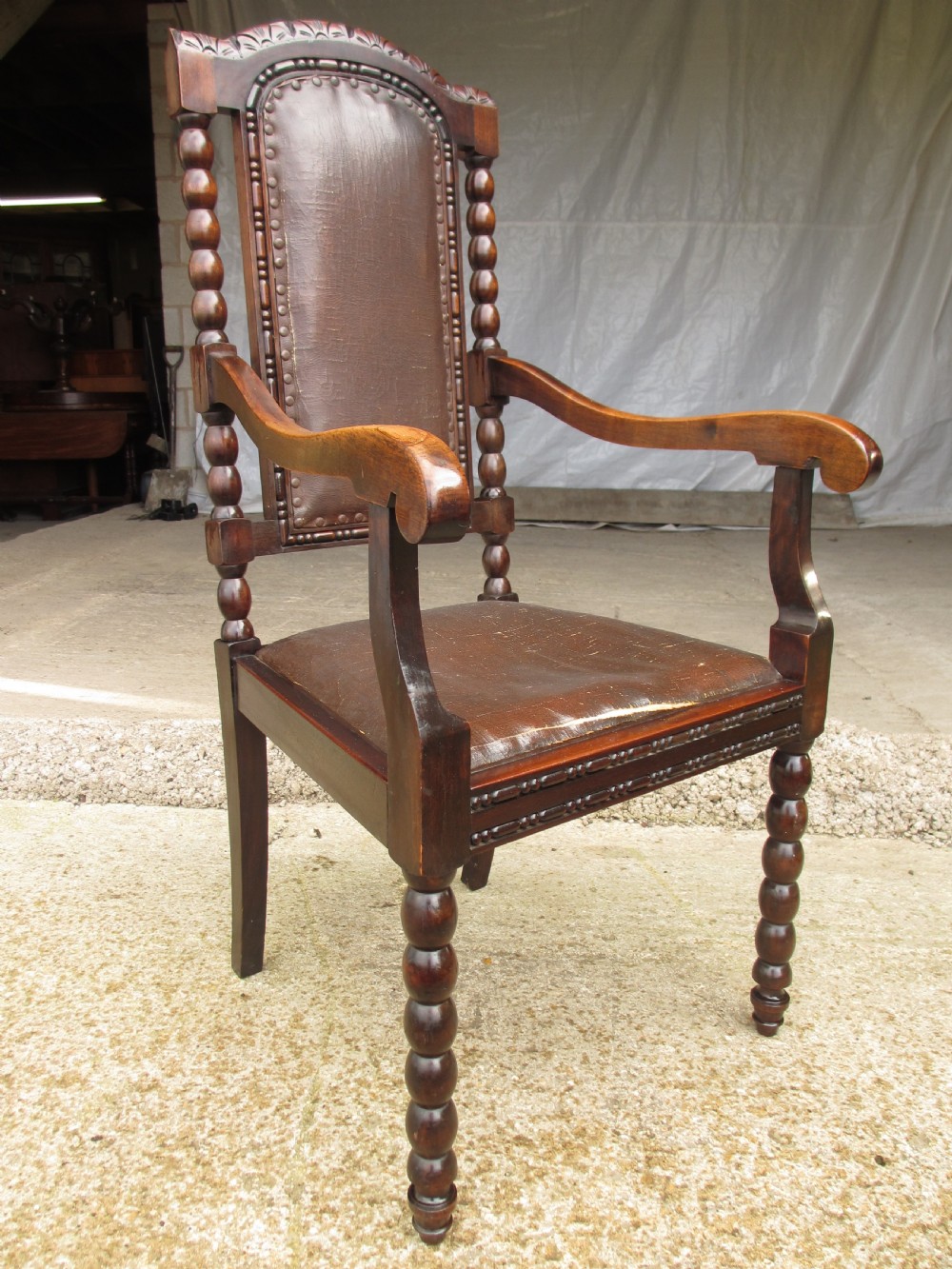 An Arts and Crafts mahogany carver armchair featuring bobbin-turned legs, curved armrests, a carved top crest, and brown studded leather upholstery. View II
