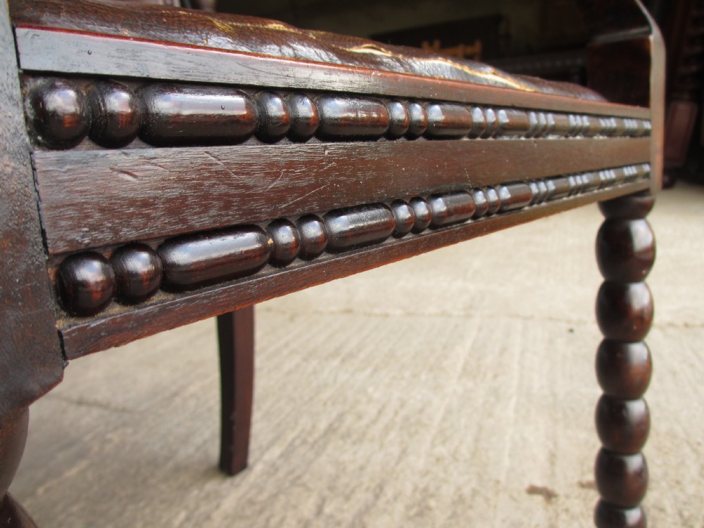 An Arts and Crafts mahogany carver armchair featuring bobbin-turned legs, curved armrests, a carved top crest, and brown studded leather upholstery. View VIII