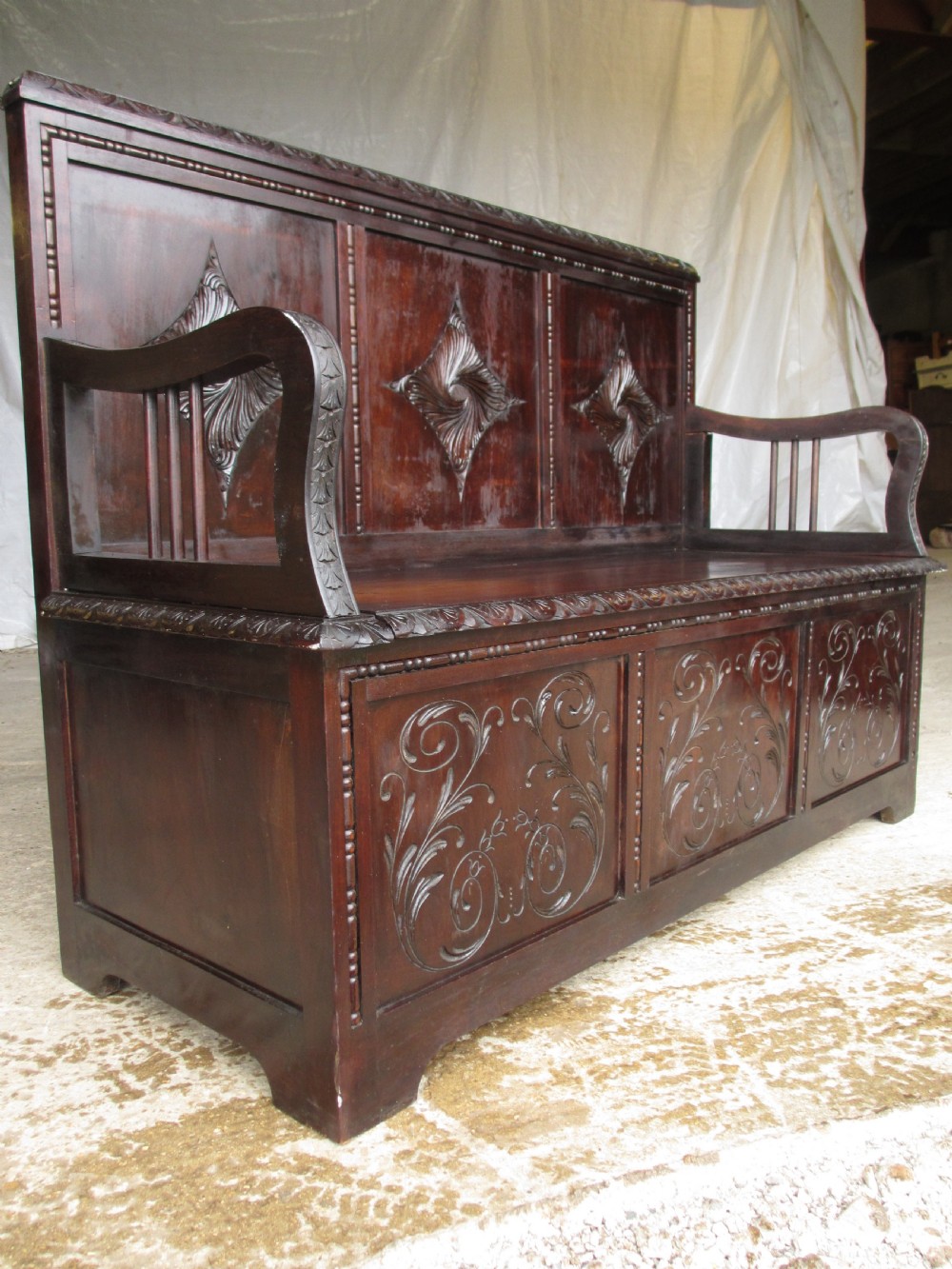 A pair of carved dark mahogany panelled box settles featuring decorative diamond back panels and scrolling foliate carved storage bases. View XV