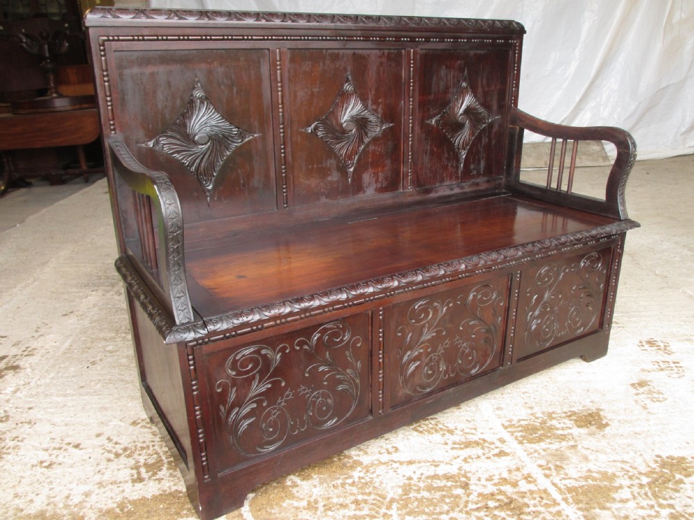 A dark mahogany panelled box settle featuring carved lozenge motifs on the backrest and foliate carving on the front panels. View II