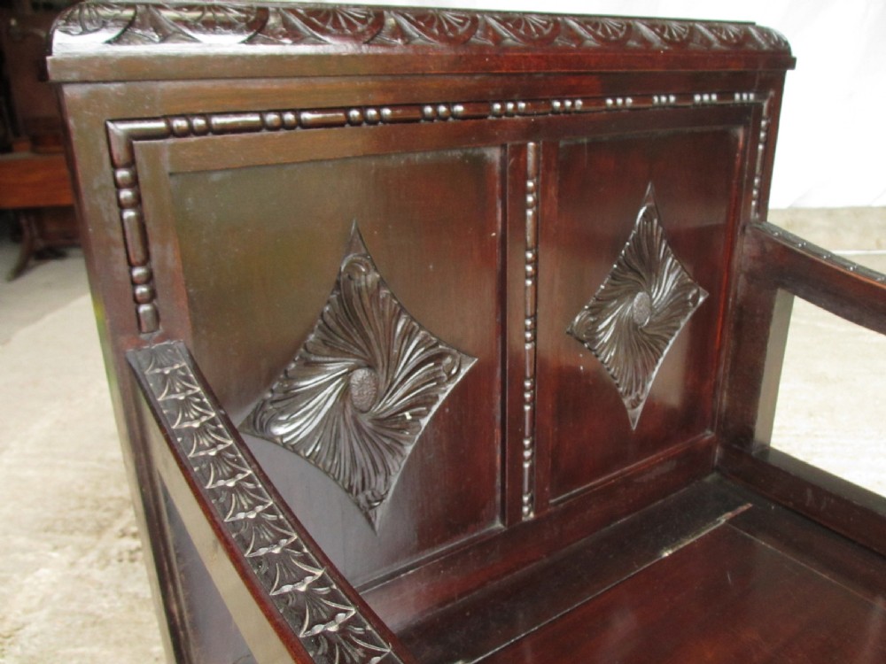 A dark brown carved mahogany box settle featuring a high panelled back with diamond motifs, curved armrests, and a hinged storage base decorated with foliate carvings. View III