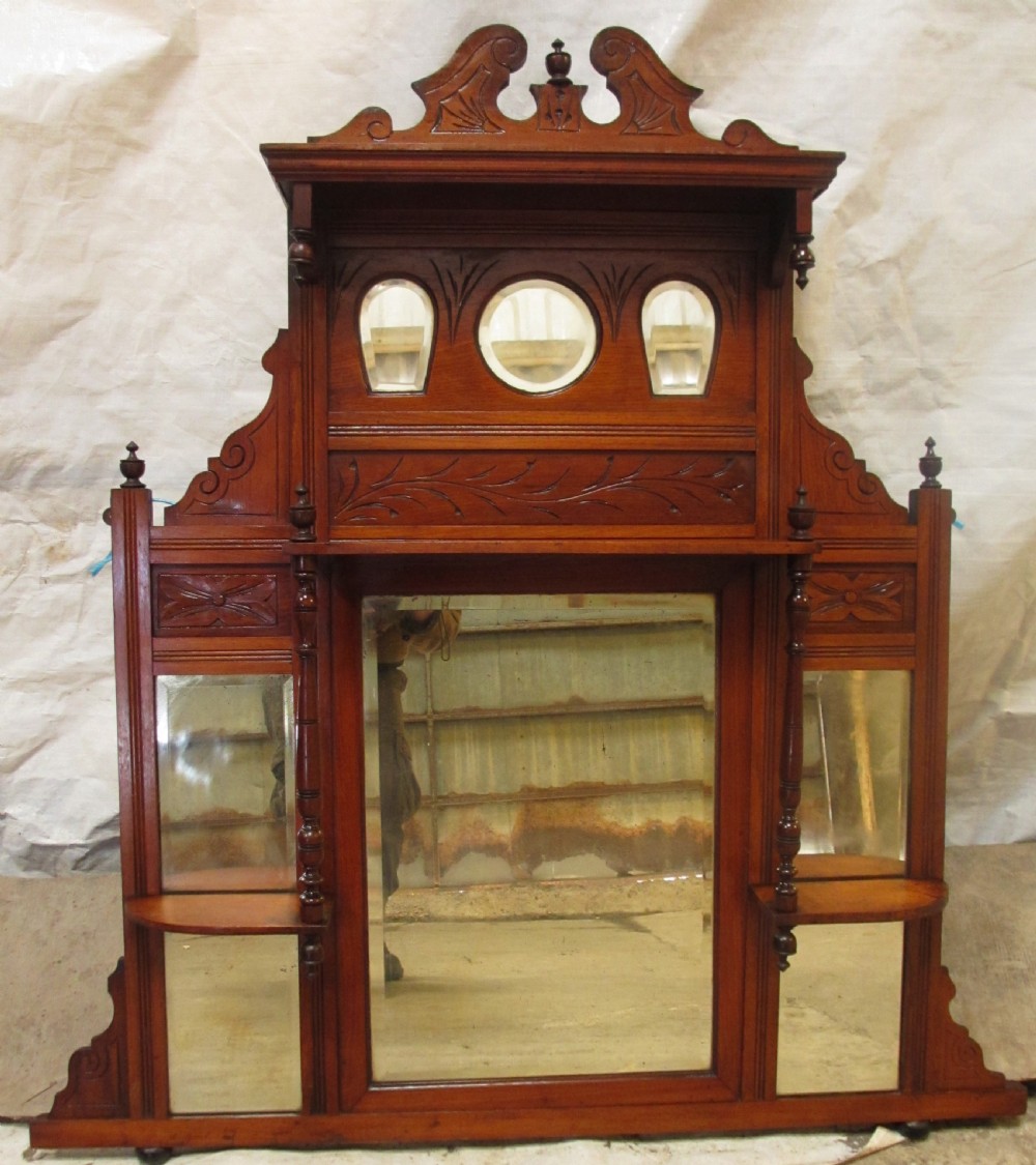 A carved mahogany Victorian overmantel mirror featuring multiple glass panels, side display shelves, turned spindle pillars, and stylised foliage carving.