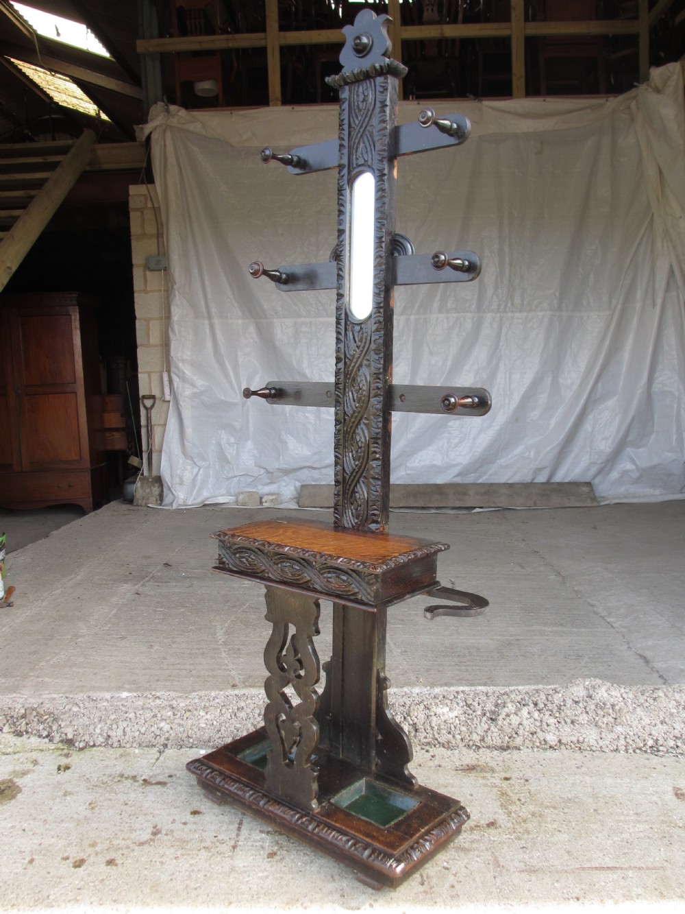 An antique Victorian dark oak hall stand featuring intricate carved patterns, turned coat pegs, a central narrow mirror, fretted base detailing, and integrated umbrella drip trays. View III