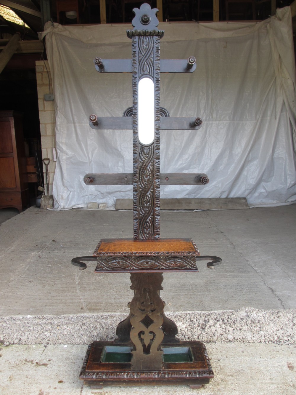 An antique Victorian dark oak hall stand featuring intricate carved patterns, turned coat pegs, a central narrow mirror, fretted base detailing, and integrated umbrella drip trays.