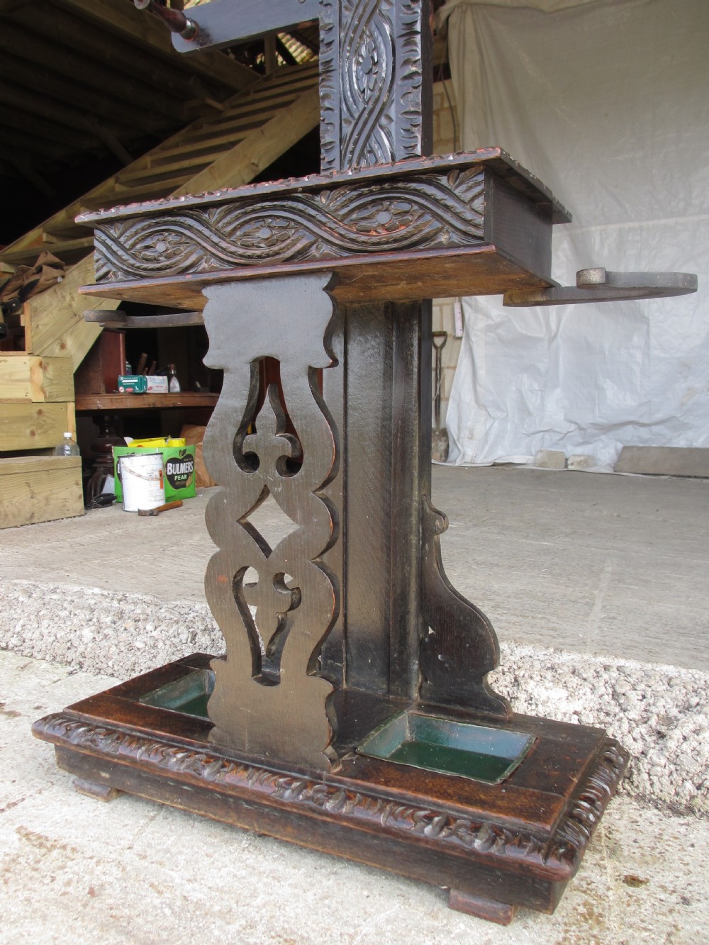 An antique Victorian dark oak hall stand featuring intricate carved patterns, turned coat pegs, a central narrow mirror, fretted base detailing, and integrated umbrella drip trays. View IV