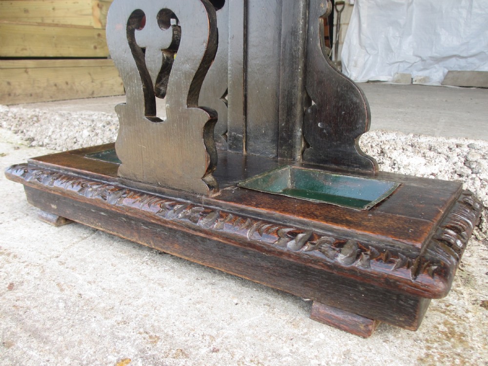 An antique Victorian dark oak hall stand featuring intricate carved patterns, turned coat pegs, a central narrow mirror, fretted base detailing, and integrated umbrella drip trays. View V