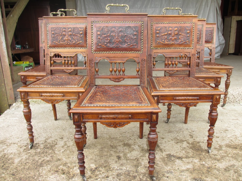 A set of Victorian carved wooden dining chairs with embossed leather backrests, studded borders, brass top handles, and turned legs with castors.