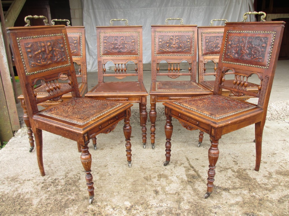 A set of Victorian carved wooden dining chairs with embossed leather backrests, studded borders, brass top handles, and turned legs with castors. View II