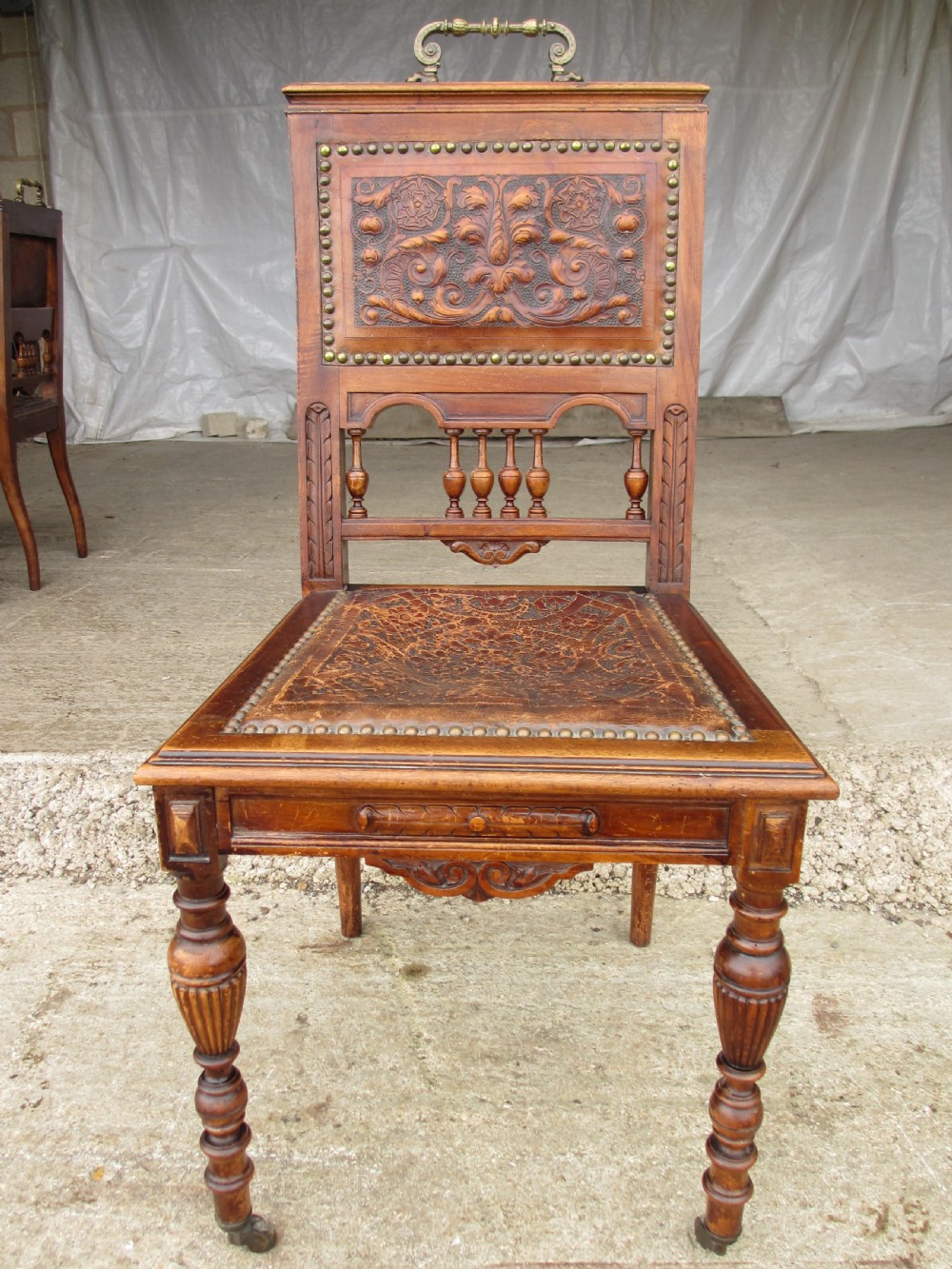 A set of Victorian carved wooden dining chairs with embossed leather backrests, studded borders, brass top handles, and turned legs with castors. View III