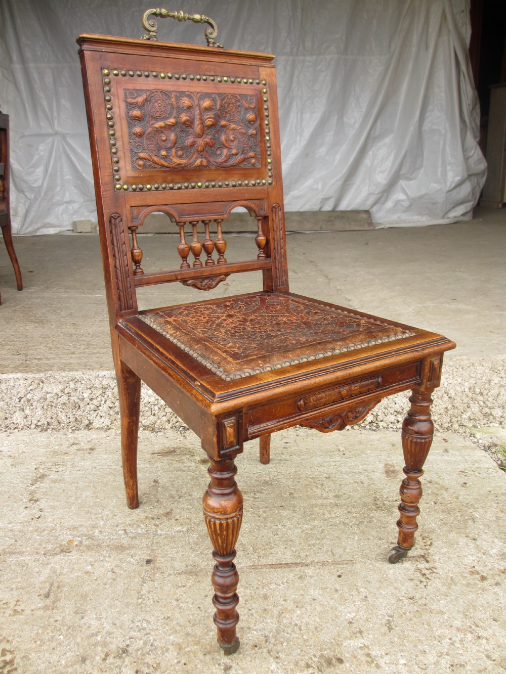 A set of Victorian carved wooden dining chairs with embossed leather backrests, studded borders, brass top handles, and turned legs with castors. View IV