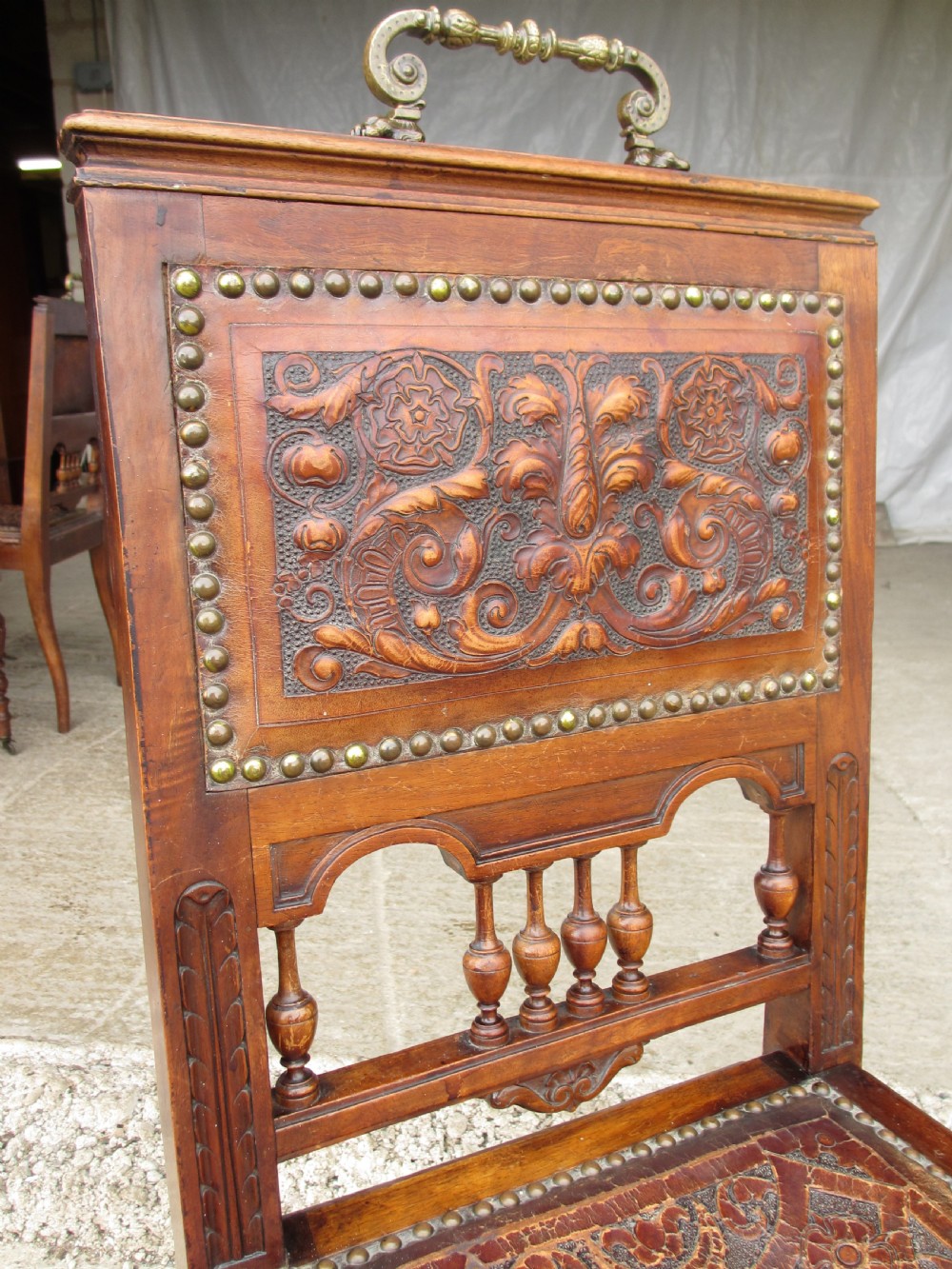 A set of Victorian carved wooden dining chairs with embossed leather backrests, studded borders, brass top handles, and turned legs with castors. View V