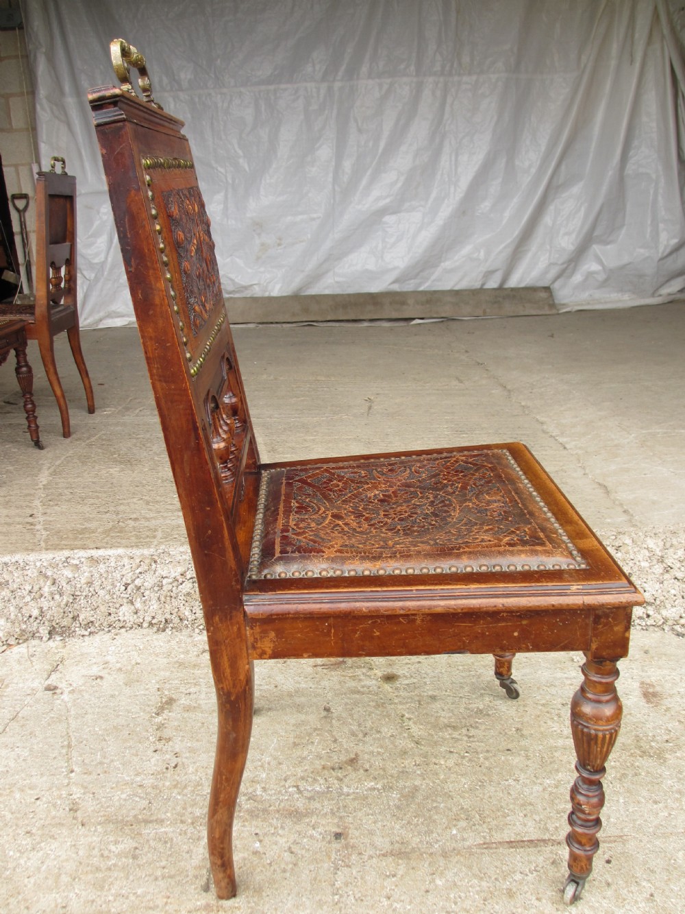 A set of Victorian carved wooden dining chairs with embossed leather backrests, studded borders, brass top handles, and turned legs with castors. View X
