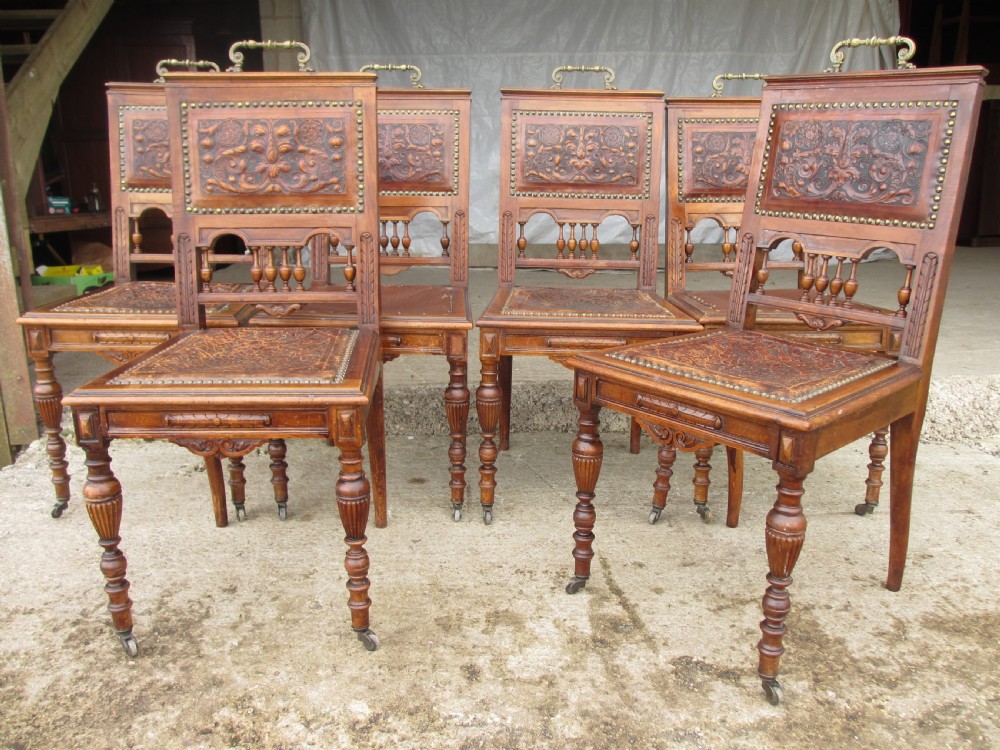 A set of Victorian carved wooden dining chairs with embossed leather backrests, studded borders, brass top handles, and turned legs with castors. View XII
