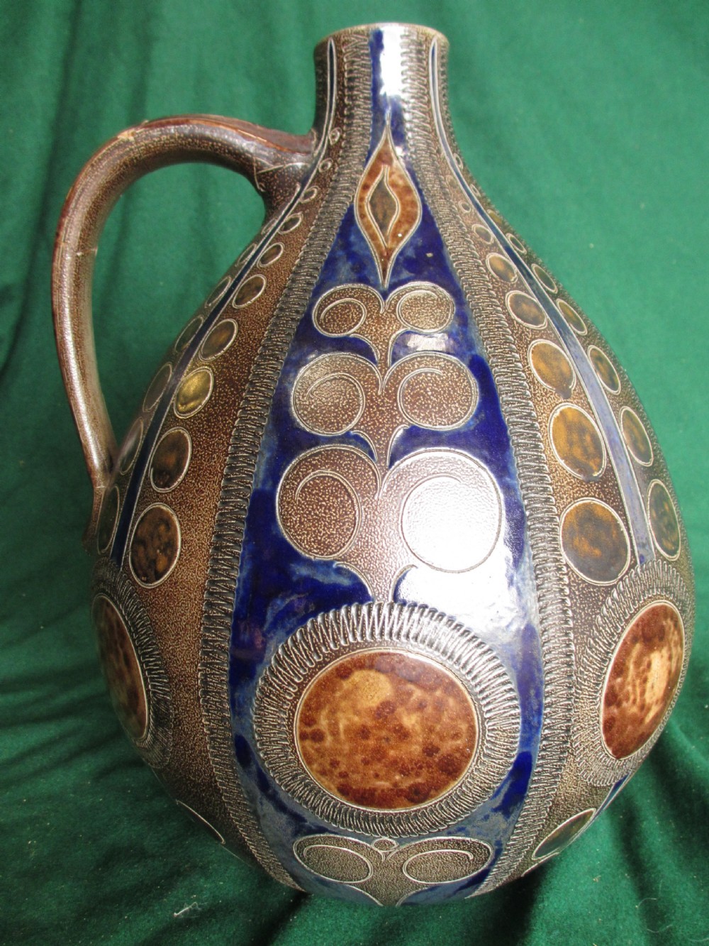 A pear-shaped Continental stoneware carafe featuring a textured surface with blue and brown glazed geometric circular motifs and a single curved handle. View III