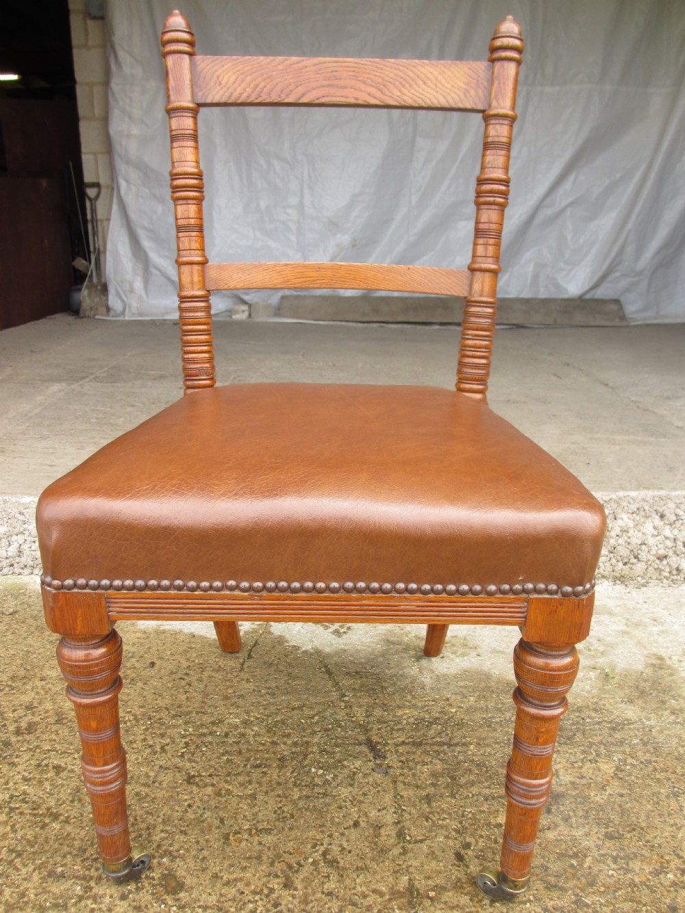 A set of four Victorian oak dining chairs featuring brown leather studded seats and turned wooden legs with brass castors. View IV