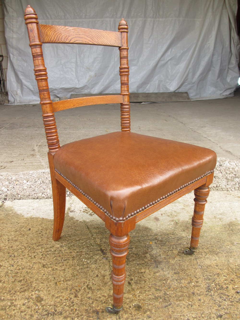 A set of four Victorian oak dining chairs featuring brown leather studded seats and turned wooden legs with brass castors. View V