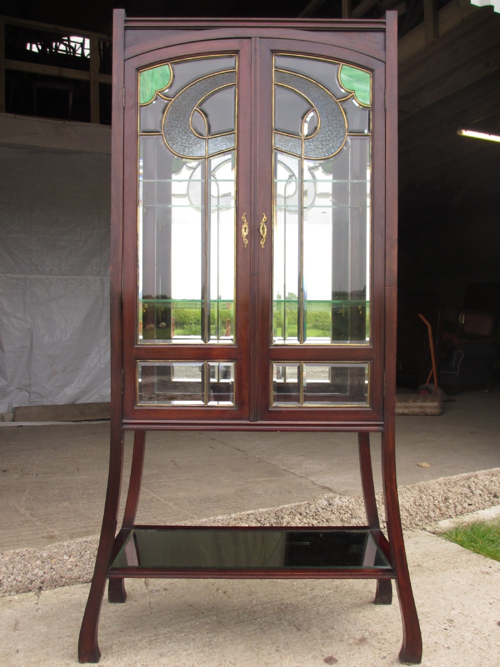 A late 19th-century mahogany Art Nouveau display cabinet featuring leaded glass doors with green accents, stylized glazing bars, interior glass shelving, and a lower open undertier.