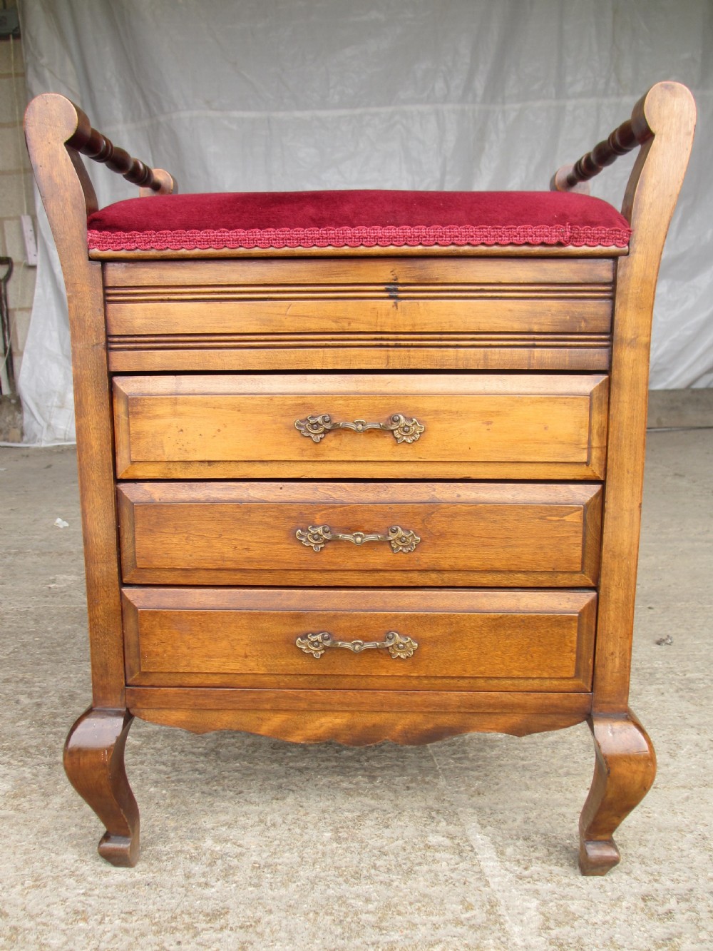 An Edwardian mahogany music stool featuring a red velvet upholstered seat, side turned handles, brass drawer pulls, and cabriole legs.