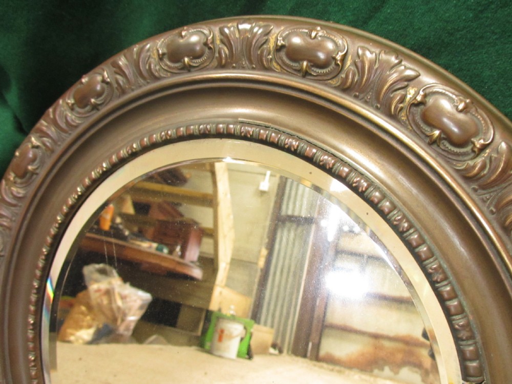 A circular Victorian brass wall mirror featuring an embossed floral patterned frame and a bevelled glass mirror plate. View IV