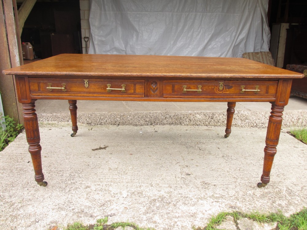 An Edwardian Arts and Crafts warm brown oak library table featuring two front drawers, a carved central floral panel, turned legs, and brass handles on castors.