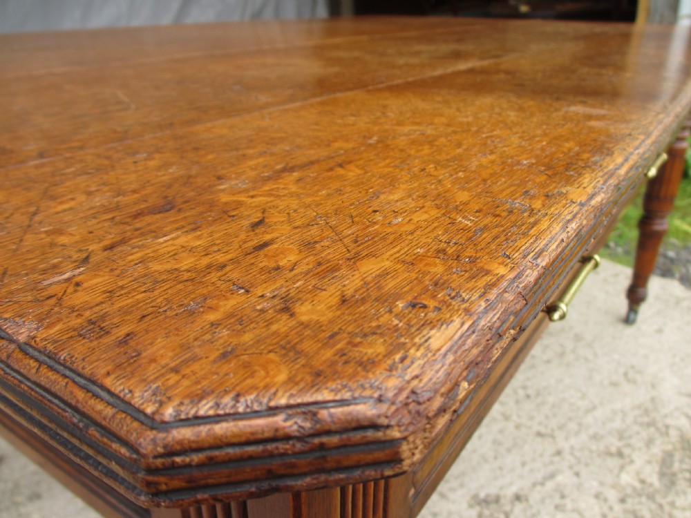 An Edwardian Arts and Crafts warm brown oak library table featuring two front drawers, a carved central floral panel, turned legs, and brass handles on castors. View II