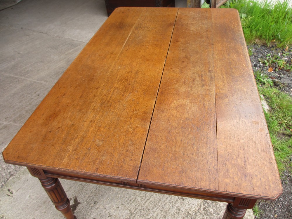 An Edwardian Arts and Crafts warm brown oak library table featuring two front drawers, a carved central floral panel, turned legs, and brass handles on castors. View III