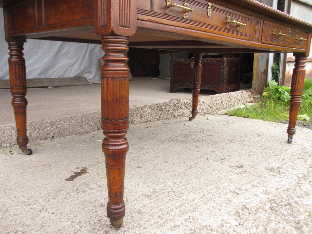 An Edwardian Arts and Crafts warm brown oak library table featuring two front drawers, a carved central floral panel, turned legs, and brass handles on castors. View IV