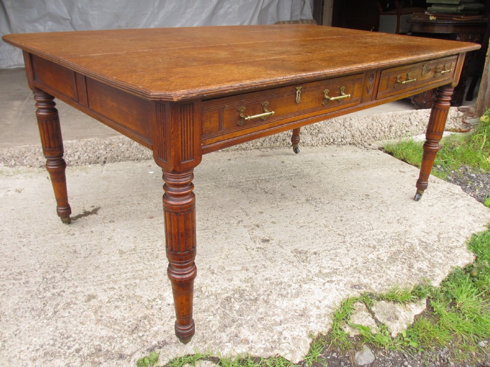 An Edwardian Arts and Crafts warm brown oak library table featuring two front drawers, a carved central floral panel, turned legs, and brass handles on castors. View VI