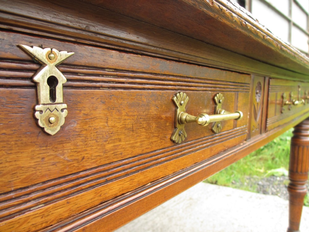 An Edwardian Arts and Crafts warm brown oak library table featuring two front drawers, a carved central floral panel, turned legs, and brass handles on castors. View VII