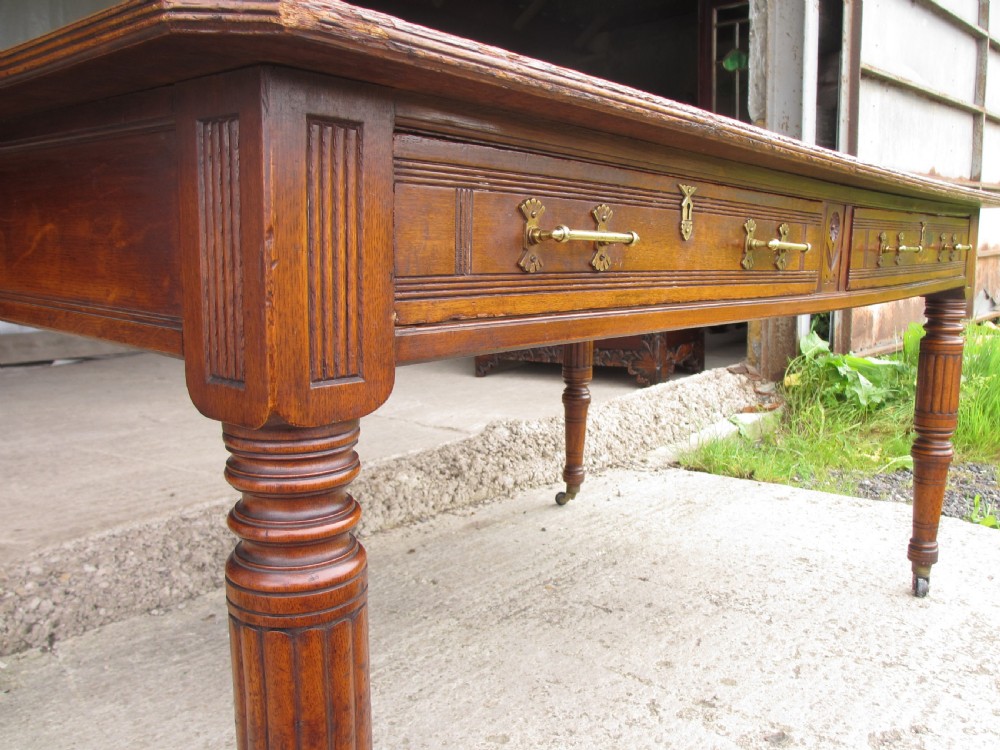 An Edwardian Arts and Crafts warm brown oak library table featuring two front drawers, a carved central floral panel, turned legs, and brass handles on castors. View VIII