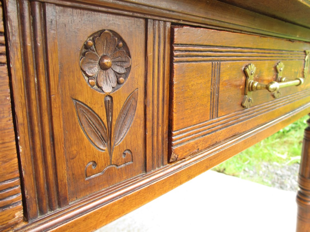 An Edwardian Arts and Crafts warm brown oak library table featuring two front drawers, a carved central floral panel, turned legs, and brass handles on castors. View IX