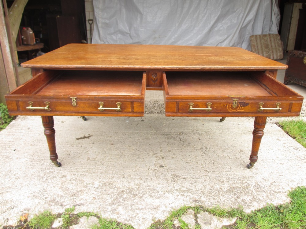 An Edwardian Arts and Crafts warm brown oak library table featuring two front drawers, a carved central floral panel, turned legs, and brass handles on castors. View X