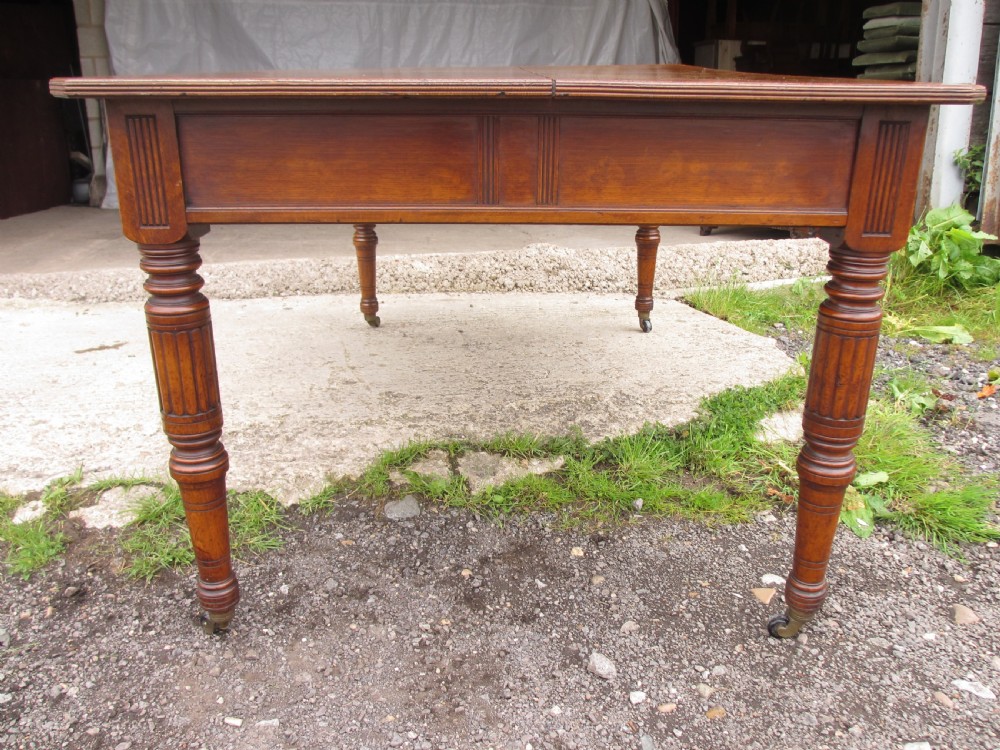 An Edwardian Arts and Crafts warm brown oak library table featuring two front drawers, a carved central floral panel, turned legs, and brass handles on castors. View XI
